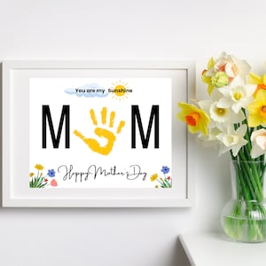 May include: A white frame with a colorful printable art print that says "You are my sunshine MOM" with a yellow handprint in the middle and the text "Happy Mother's Day" at the bottom. The frame is sitting on a white surface with a vase of yellow daffodils in the background.