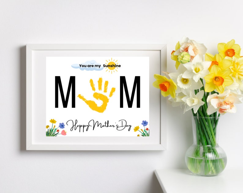 May include: A white frame with a colorful printable art print that says "You are my sunshine MOM" with a yellow handprint in the middle and the text "Happy Mother's Day" at the bottom. The frame is sitting on a white surface with a vase of yellow daffodils in the background.