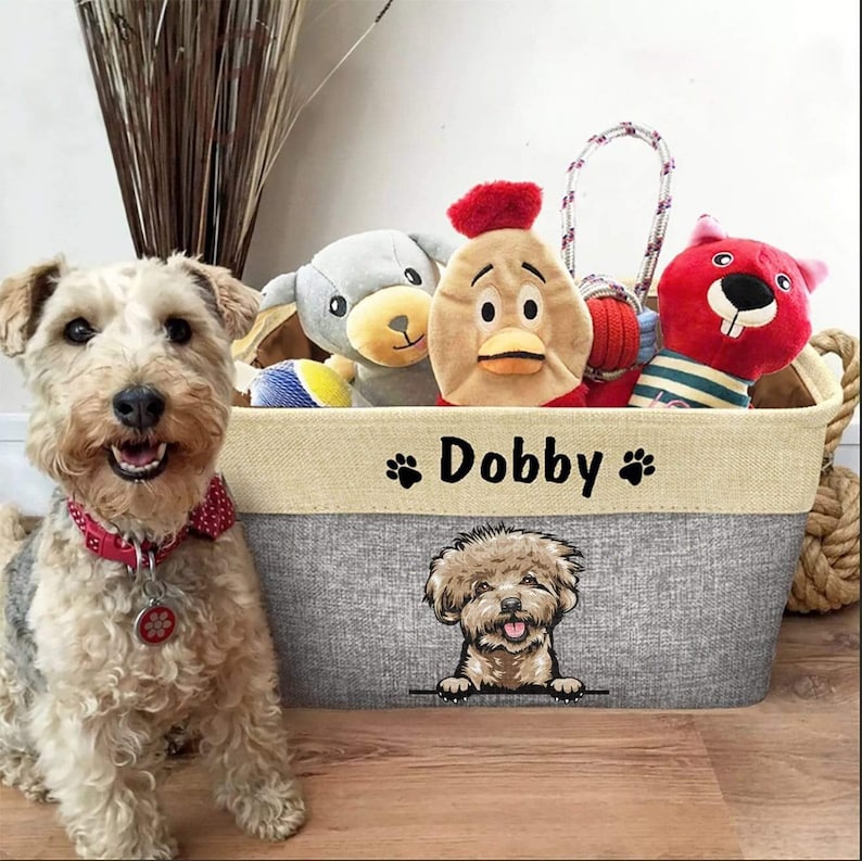 Pode incluir: Uma cesta de armazenamento cinza e bege com o nome "Dobby" bordado na frente. A cesta est&aacute; cheia de brinquedos para c&atilde;es, incluindo um brinquedo de frango vermelho e branco, um brinquedo de pel&uacute;cia cinza e branco e um brinquedo de corda vermelho e branco. Um c&atilde;o marrom e branco est&aacute; sentado na frente da cesta.