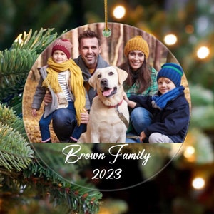 May include: A round glass Christmas bauble featuring a family photo of four people and a dog. The family are wearing winter clothing and are standing in a forest. The text "Brown Family 2023" is printed on the bauble.