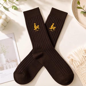 May include: A pair of dark brown ribbed crew socks featuring a small yellow graphic of two figures in a sexual position with the letters A and J separated by a heart symbol embroidered on the upper portion of each sock.