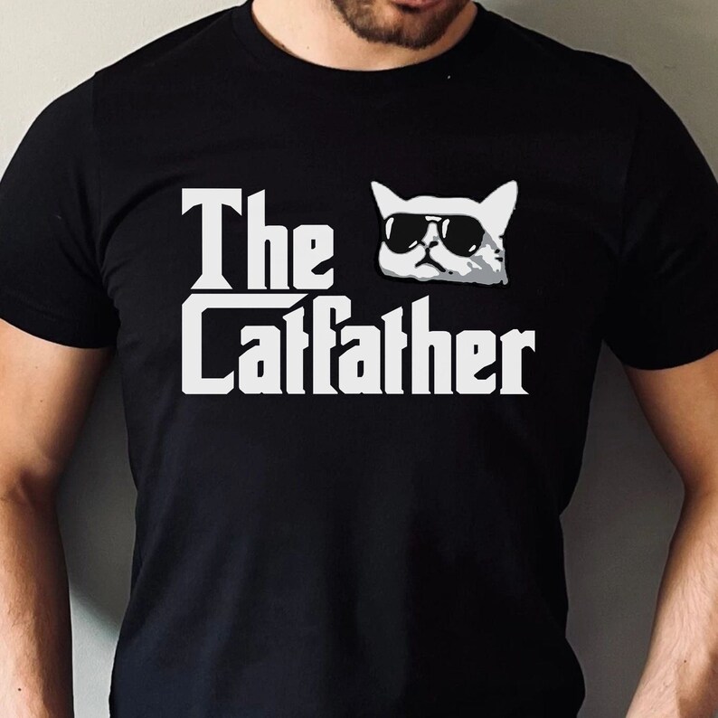 May include: A black t-shirt with a white graphic of a cat wearing sunglasses and the text "The Catfather".