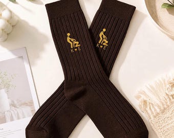 May include: A pair of dark brown ribbed crew socks featuring an embroidered gold graphic of two stick figures in an intimate pose, positioned above the letters K and E separated by a small heart symbol, centred on the upper part of each sock.