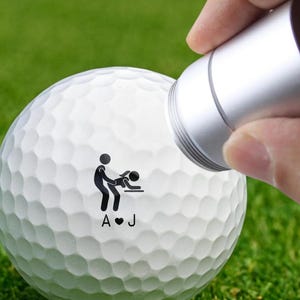 May include: A white golf ball with a black graphic of two stick figures in a sexual position, with the initials "A" and "J" and a heart symbol. A silver tool is held near the ball.