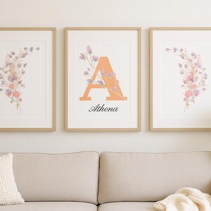 Puede incluir: Tres impresiones artísticas florales enmarcadas. Dos impresiones presentan un arreglo floral en acuarela en tonos rosa, morado y verde. La impresión central muestra la letra "A" en naranja, con el nombre "Athena" debajo. Los marcos son de color marrón claro.