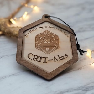 May include: A wooden Christmas ornament shaped like a hexagon with a 20-sided die in the centre and the text "It's Beginning to Look a Lot Like CRIT-Mas".