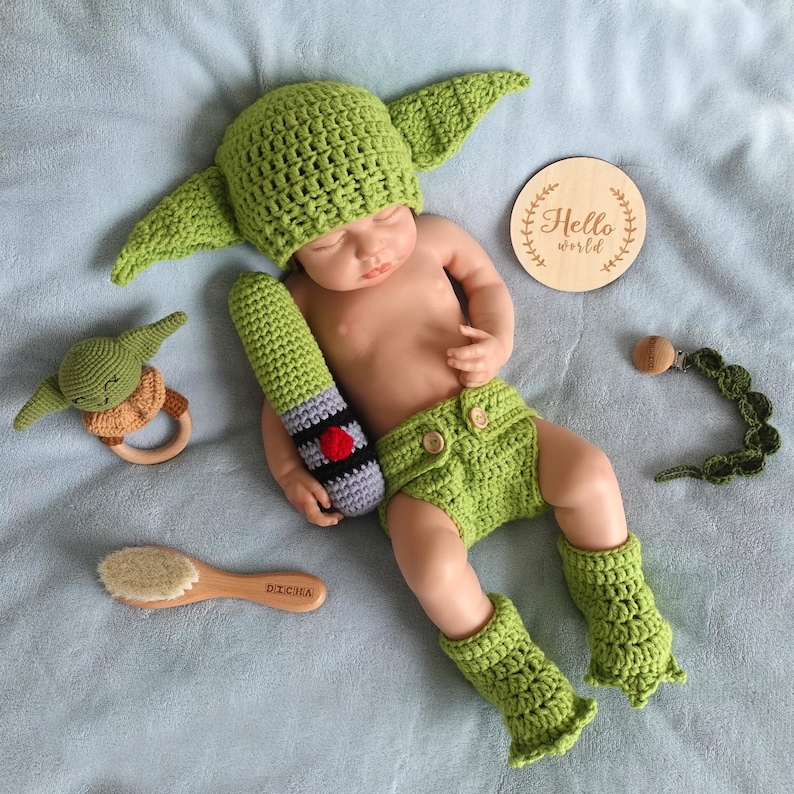 Op de afbeelding: Een baby in een groen gehaakt Yoda-kostuum, inclusief een hoed met grote oren, een korte broek en beenwarmers. Accessoires: een gehaakte lichtzwaard, een houten bijtring, een fopspeenclip, een houten borstel en een houten schijf met "Hello world".