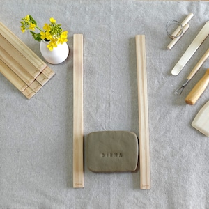 May include: Two wooden dowels, each 10 inches long, are placed vertically on a light beige fabric surface. A rectangular piece of brown clay with the word "DIGHA" embossed on it is placed between the dowels.