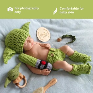 Op de afbeelding: Een pasgeboren baby in een groen gehaakt Yoda-thema-outfit, inclusief een hoed, shorts en beenwarmers. Accessoires zijn onder meer een lichtzwaard, een houten bord met "Hello world" en een rammelaar. De tekst in de afbeelding zegt "For photography only" en "Comfortable for baby skin".