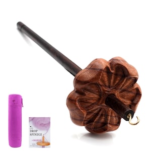 May include: A dark wood drop spindle with a carved, flower-shaped whorl. The spindle has a long, slender shaft and a metal hook. A purple carrying case and a manual are also in the image.