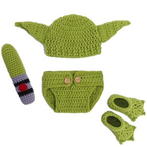 Op de afbeelding: Een groen gehaakt baby-outfit dat lijkt op een populair personage, inclusief een hoed met grote oren, een luierbroekje met houten knopen en babyslofjes. Een gehaakte lichtzwaard is ook inbegrepen. De set is ontworpen voor baby's.