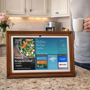 Puede incluir: Una pantalla digital con marco de madera que muestra una receta de ensalada de feta y rúcula, información meteorológica y un calendario. La pantalla muestra la hora, la temperatura e iconos de Prime Video y YouTube. Una persona sostiene una taza blanca en el fondo.