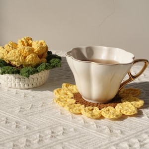 May include: A white teacup with a gold rim and handle sits on a crocheted sunflower coaster. The cup is filled with tea and there is a crocheted flower pot with yellow flowers in the background.