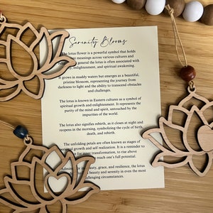 May include: Two wooden lotus flower ornaments with a brown string and wooden beads. The ornaments are hanging from the string. The text on the card reads "Serenity Blooms". The lotus flower is a powerful symbol that holds many meanings across various cultures and in general the lotus is often associated with purity, enlightenment, and spiritual awakening.