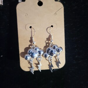 May include: A pair of silver-toned drop earrings featuring a cloud-shaped charm with a polished metallic finish. Each cloud has two small lightning bolt charms dangling from the bottom, attached to a standard silver-toned fishhook ear wire.