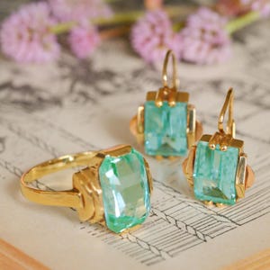 May include: A set of vintage jewelry featuring a ring and earrings with light green gemstone accents. The gold-toned pieces have a delicate, Art Deco-inspired design.
