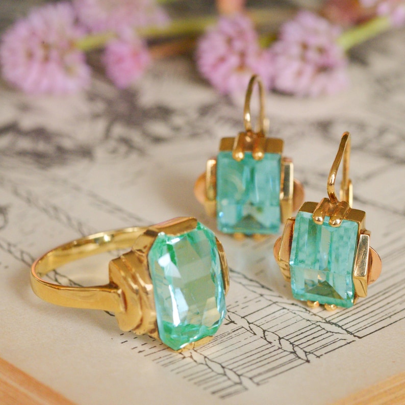 May include: A set of vintage jewelry featuring a ring and earrings with light green gemstone accents. The gold-toned pieces have a delicate, Art Deco-inspired design.