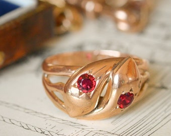May include: A gold snake ring with two red ruby stones set in the eyes. The ring is detailed with a textured snake body.