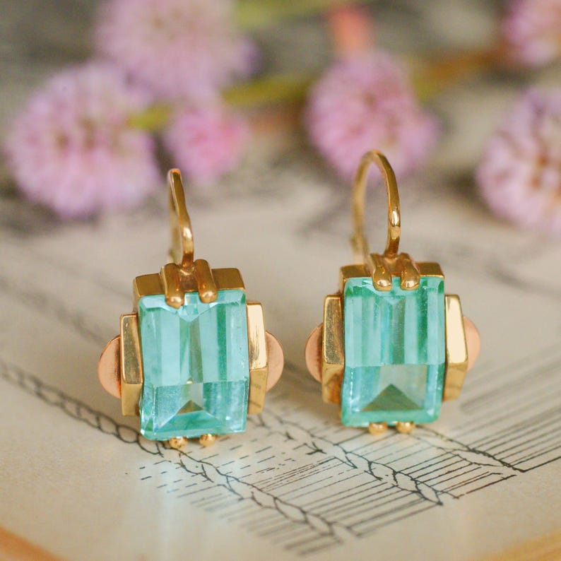 May include: A pair of gold earrings with light blue square-shaped gemstones. The earrings have a geometric design and are accented with rose gold.