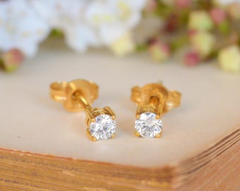 May include: A pair of gold stud earrings with a round, clear gemstone. The earrings are set on a light-colored surface with a blurred floral background. The earrings are a classic jewelry item.