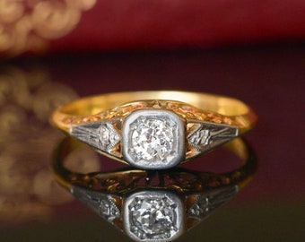 May include: A vintage engagement ring with a delicate filigree design. The ring is made of gold and features a round, brilliant-cut diamond in a square setting. Two smaller diamonds are set on either side of the center stone.