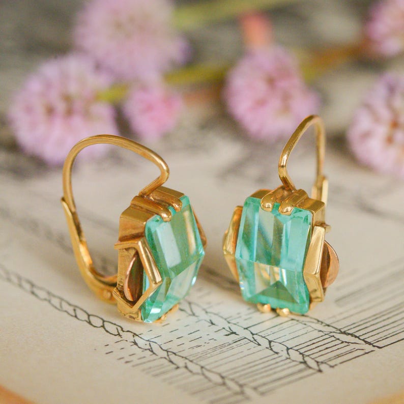 May include: A pair of gold earrings with light green gemstone accents. The earrings have a simple, elegant design and are perfect for everyday wear.