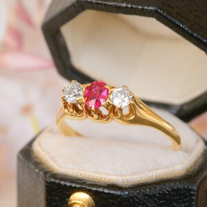 May include: A gold ring featuring a red ruby and two white diamonds. The ring is displayed in a black ring box.