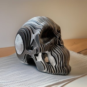 May include: A layered metal skull sculpture with a detailed, intricate design. The skull is made of multiple layers of metal, creating a 3D effect. The sculpture is painted in a silver colour.
