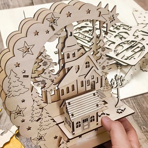 Op de afbeelding: Een houten kerststal met een kerk, huizen en bomen. De stal is gemaakt van lasergesneden hout en is ontworpen om in elkaar te zetten.