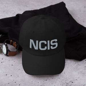 Peut inclure: Casquette de baseball noire avec un logo "NCIS" brodé en blanc.