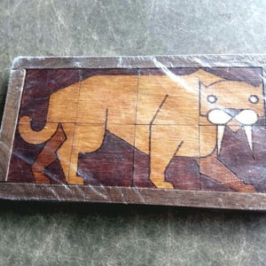 May include: Wooden puzzle featuring a saber-toothed tiger. The puzzle is made of various shades of brown wood, with the tiger in a lighter tone against a darker background. The puzzle is rectangular and appears to be new, still in its original packaging.