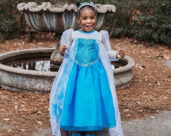 May include: A young girl wearing a blue and white princess costume with a sparkly bodice and a white cape. She is standing in front of a stone fountain.