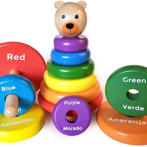 Könnte beinhalten: Ein Holz-Stapelspielzeug mit einem Bärenkopf oben und bunten Ringen. Die Ringe sind mit den Farben Rot, Blau, Amarillo, Lila, Grün und Naranja beschriftet.
