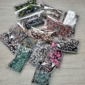 May include: A collection of small, decorative polymer clay pieces in various colors and patterns, packaged in clear plastic bags. Designs include skulls, smiley faces, and floral patterns. The pieces are scattered on a light wood-grain surface.