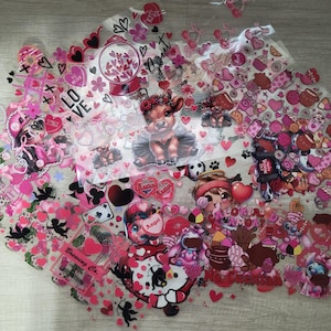 May include: Assortment of pink and red Valentine's Day themed stickers and embellishments. The stickers feature hearts, angels, a cow, a cartoon character, and the text "Calorias de Amor? No. Cuentan".