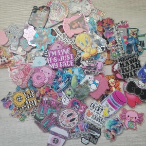 May include: A collection of colorful, glittery resin flatback embellishments featuring various cartoon characters, animals, and phrases like "I'm fine it's just my face" and "Somebody's Hawk Tuah Wif".