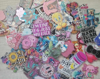 May include: A collection of colorful, glittery resin flatback embellishments featuring various cartoon characters, animals, and phrases like "I'm fine it's just my face" and "Somebody's Hawk Tuah Wif".