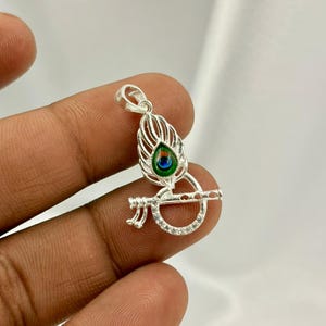 Op de afbeelding: Een zilveren hanger met een pauwenveer- en fluitontwerp. De veer heeft groene en blauwe emaille details. De fluit is versierd met kleine, heldere stenen. De hanger wordt door een hand vastgehouden, wat het ingewikkelde ontwerp laat zien.