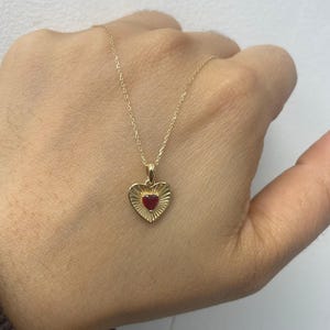 Könnte beinhalten: Goldfarbene Halskette mit herzförmigem Anhänger und rotem Mittelstein. Der Anhänger hat ein geriffeltes Design. Die Halskette wird vor einem neutralen Hintergrund präsentiert.