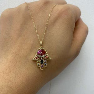 May include: A gold necklace with a Hamsa hand pendant. The pendant is decorated with a variety of colourful gemstones, including red, blue, and purple. The necklace is held up by a hand.