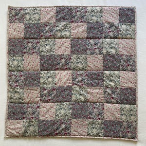 Peut inclure: Une courtepointe carrée en patchwork avec un motif en damier. La courtepointe présente des carrés alternés de tissu floral dans des tons de rose, de gris et de vert. La courtepointe est probablement en coton et a une texture matelassée.