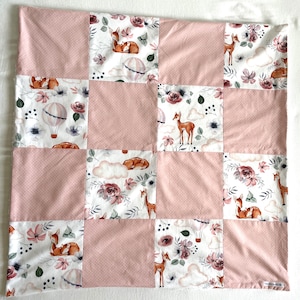 Peut inclure: Une courtepointe en patchwork avec des carrés roses à pois et des motifs floraux alternés. Les carrés floraux présentent des cerfs, des montgolfières et des fleurs dans des tons de marron, rose et blanc. La courtepointe est de forme carrée.
