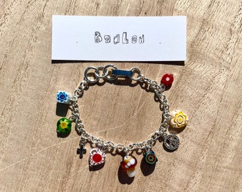May include: A silver charm bracelet with a variety of colorful glass charms, including a flower, a mushroom, a heart, and a peace sign. The bracelet has a clasp and is on a wooden surface.