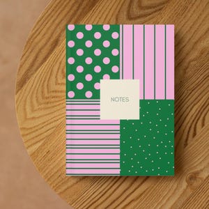 Puede incluir: Un cuaderno con un diseño de patchwork colorido. La portada presenta patrones verdes y rosas, incluyendo lunares, rayas y pequeños puntos. Un cuadrado color crema en el centro dice "NOTES". El cuaderno está sobre una mesa de madera.