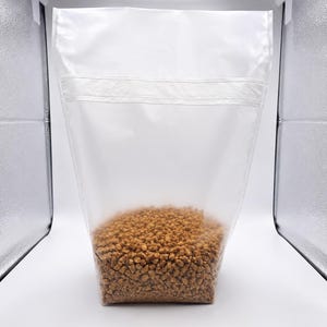 May include: A clear plastic bag filled with brown, granular material. The bag is sealed at the top with a horizontal seam. The contents appear to be pet food or a similar product. The background is a white studio setting.