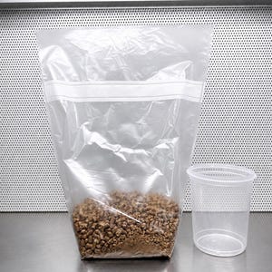 May include: A clear plastic bag filled with brown substrate, sealed with a white strip, and a clear plastic cup with perforated sides. The items are on a stainless steel surface.