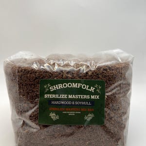 May include: A clear plastic bag filled with brown material. A green label on the bag reads "SHROOMFOLK STERILIZE MASTERS MIX HARDWOOD & SOYHULL STERILIZE MASTERS MIX BAG USED FOR WOOD LOVING SPECIES."