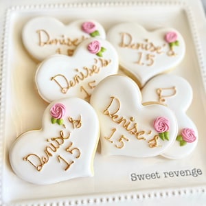 May include: A white platter with five heart-shaped cookies decorated with pink roses and gold lettering. The cookies say "Denise's 15". The platter has the words "Sweet revenge" printed on the bottom.