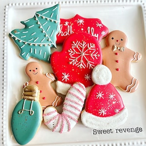 May include: A set of nine Christmas-themed sugar cookies decorated with icing and sprinkles. The cookies include a Christmas tree, a mitten, a gingerbread person, a candy cane, a light bulb, and a Santa hat. The cookies are arranged on a white plate with the words "Sweet revenge" written on the bottom.