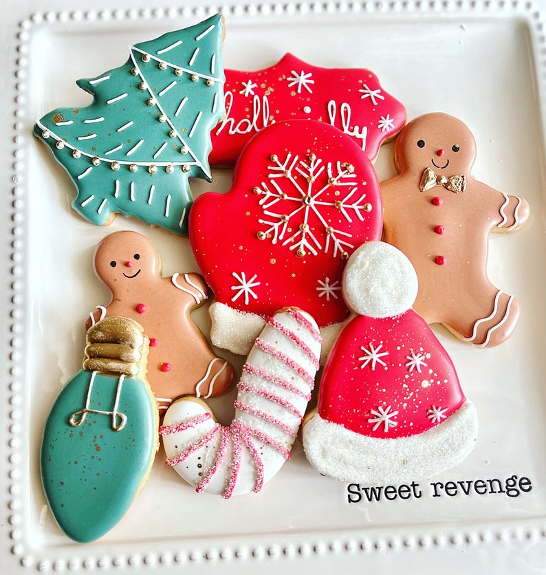 May include: A set of nine Christmas-themed sugar cookies decorated with icing and sprinkles. The cookies include a Christmas tree, a mitten, a gingerbread person, a candy cane, a light bulb, and a Santa hat. The cookies are arranged on a white plate with the words "Sweet revenge" written on the bottom.